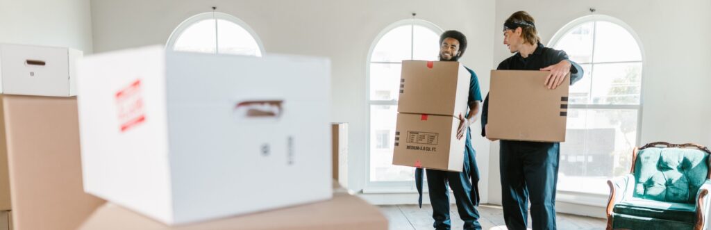Zwei gutgelaunte Umzugsmitarbeiter tragen Kartons im Raum eines Altbaus mit großen Fenstern