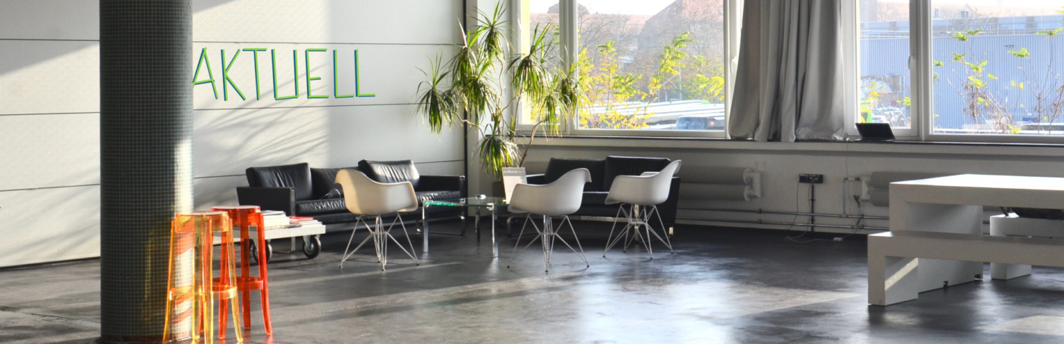 Großer, heller Raum mit großen Fenstern, Sofas, Eames-Wire-Chairs, zwei Sitzbänken mit Tisch, einer großen Pflanze und zwei farbigen Barhockern