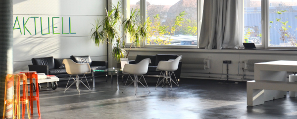 Großer, heller Raum mit großen Fenstern, Sofas, Eames-Wire-Chairs, zwei Sitzbänken mit Tisch, einer großen Pflanze und zwei farbigen Barhockern