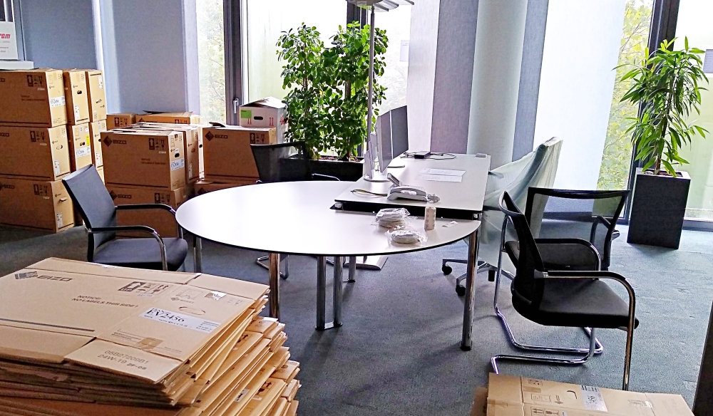 In einem Open Space Büro, in dem ein Leitungsarbeitsplatz mit zwei Bildschirmen und Besprechungsanbau steht, befinden sich viele leere und volle Umzugskartons