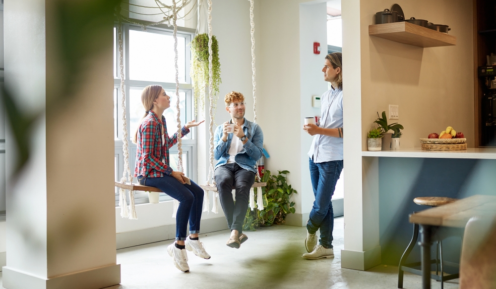 An einem Meeting Point mit Küche, Pflanzen und zwei Schaukeln unterhalten sich informell drei jüngere Mitarbeitende